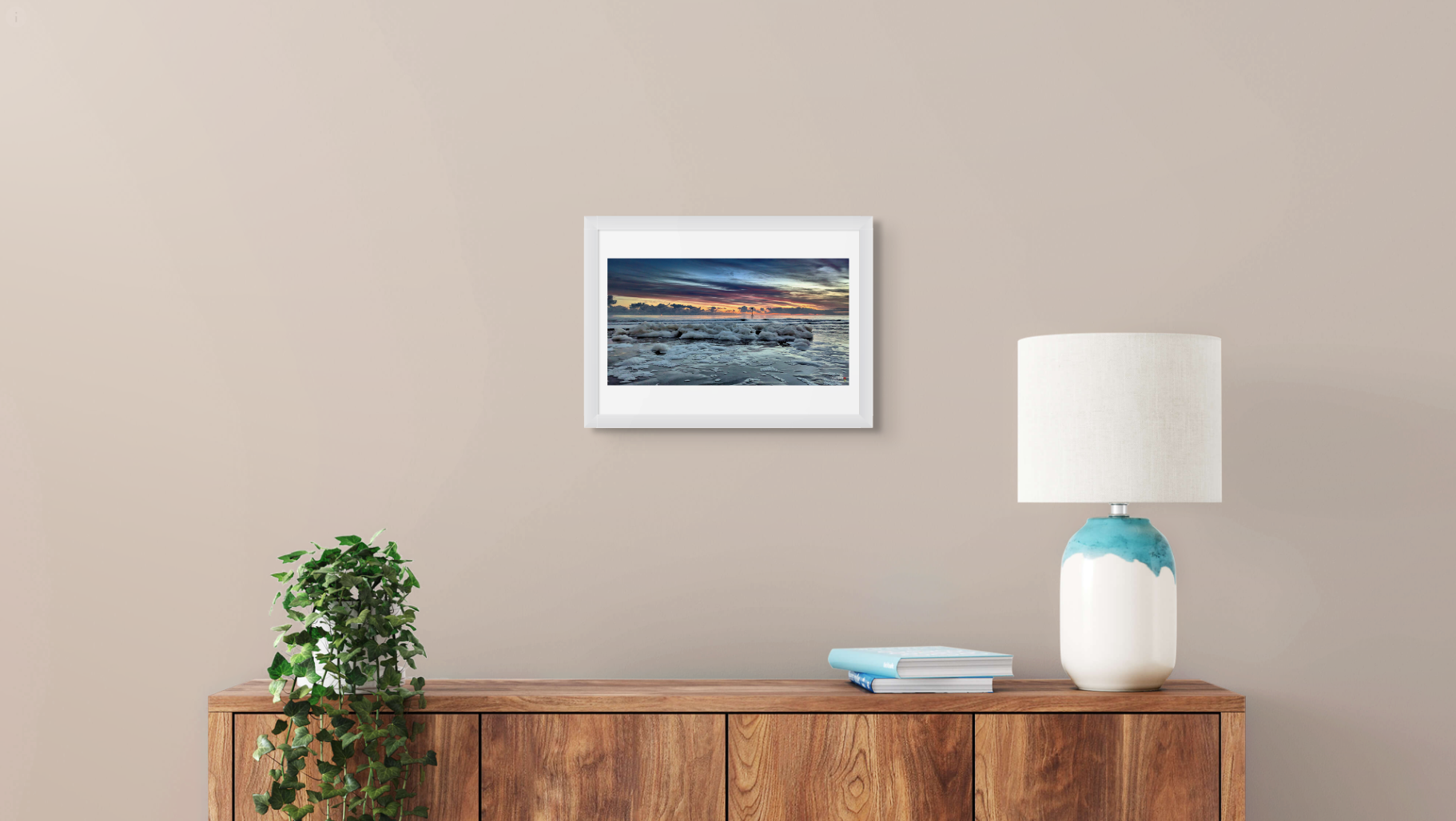 Framed artwork on a wall above a wooden sideboard with a lamp and plant.