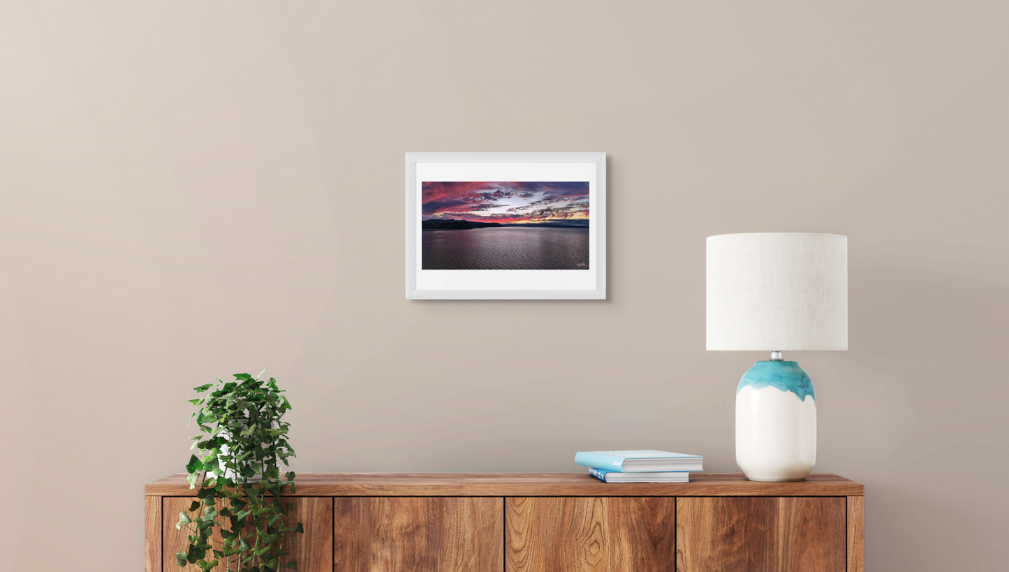 Framed artwork on a wall above a wooden sideboard with a lamp and plant.