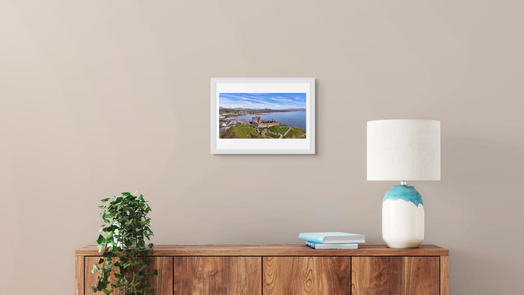 Framed landscape print on a wall above a wooden sideboard with a lamp and plant.