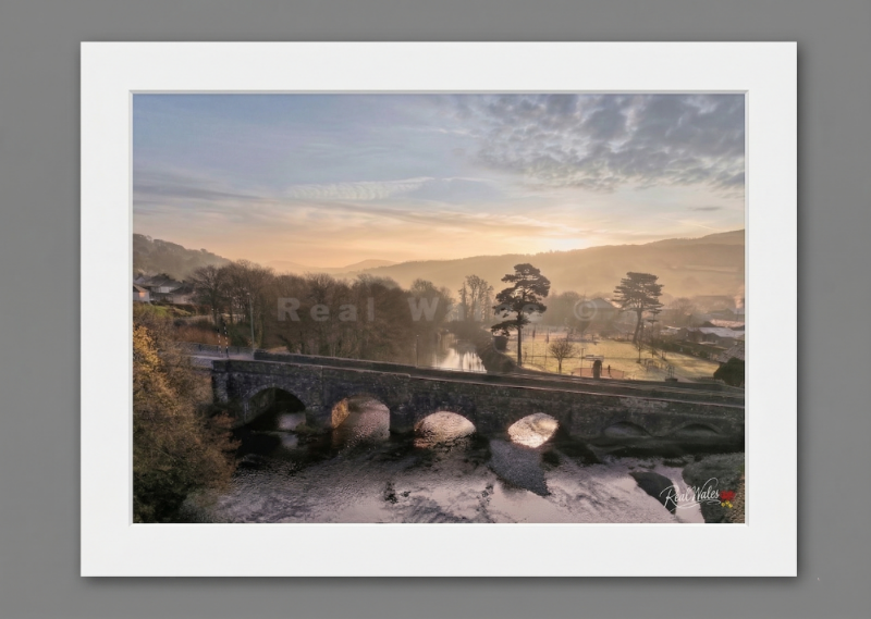 The sturdy, multi-arched span of Dolgellauâ€™s historic stone bridge anchors the town across the gentle flow of the River Wnion, forging an enduring path through the frost-touched valley. Long celebrated for its striking mountain enclosure and the quiet majesty of its waterways, this market town awakens with a sense of peaceful, earthbound permanence. The spreading, golden warmth of the morning sun and the soft, lifting river mist offer a luminous, waking contrast to the cold, weather-beaten weight of the d