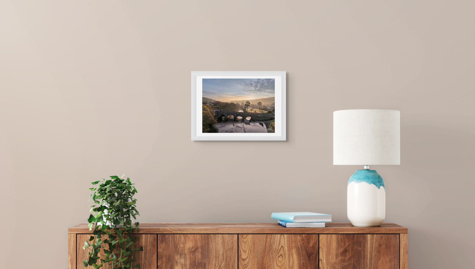 Framed artwork on a wall above a wooden console table with a lamp and books.