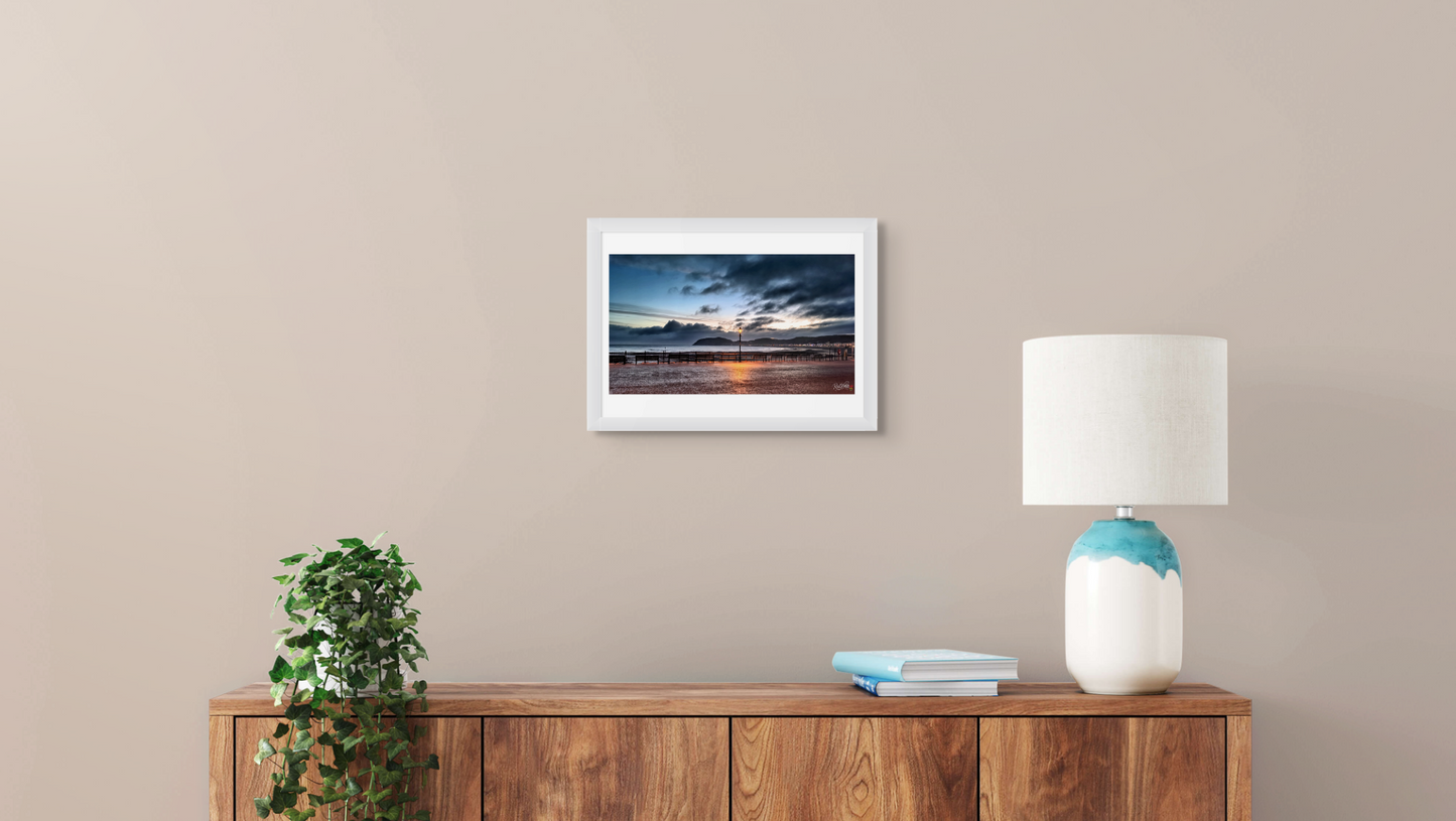 Framed artwork on a wall above a wooden console table with a lamp and books.