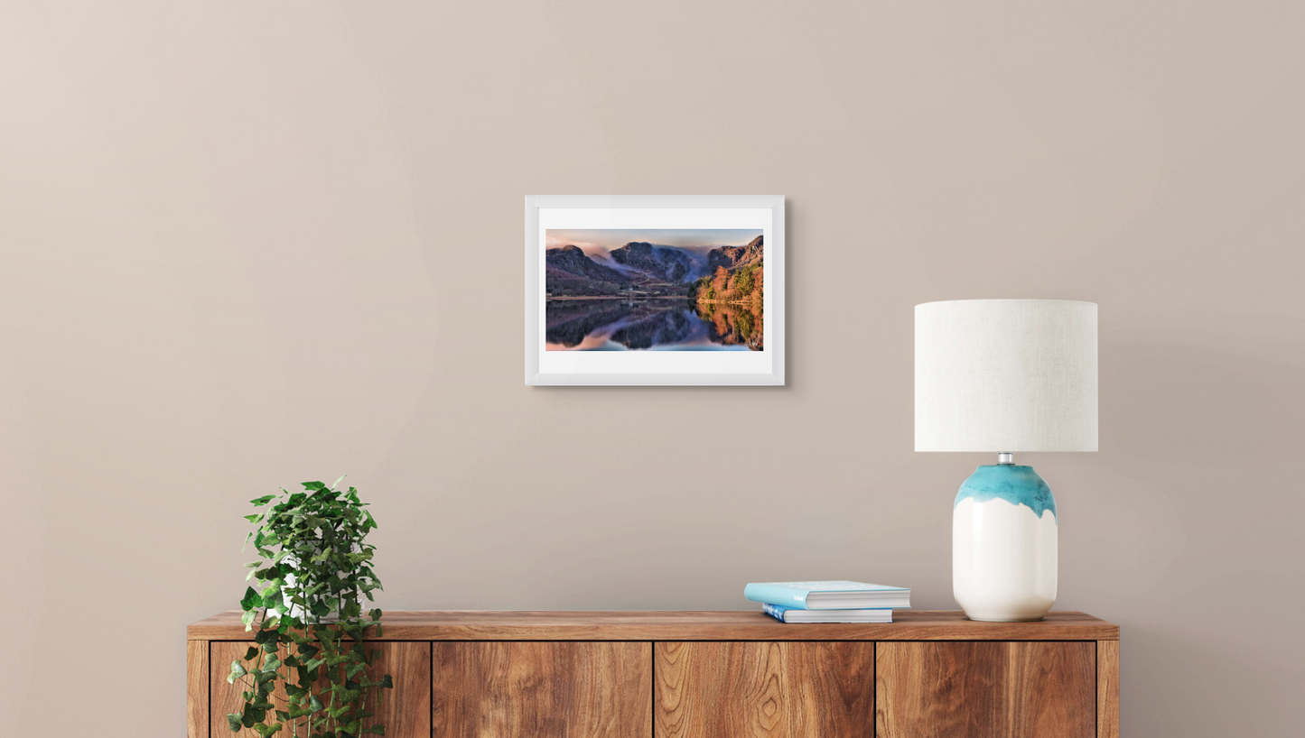 Framed landscape print on a wall above a wooden sideboard with a lamp and plant.