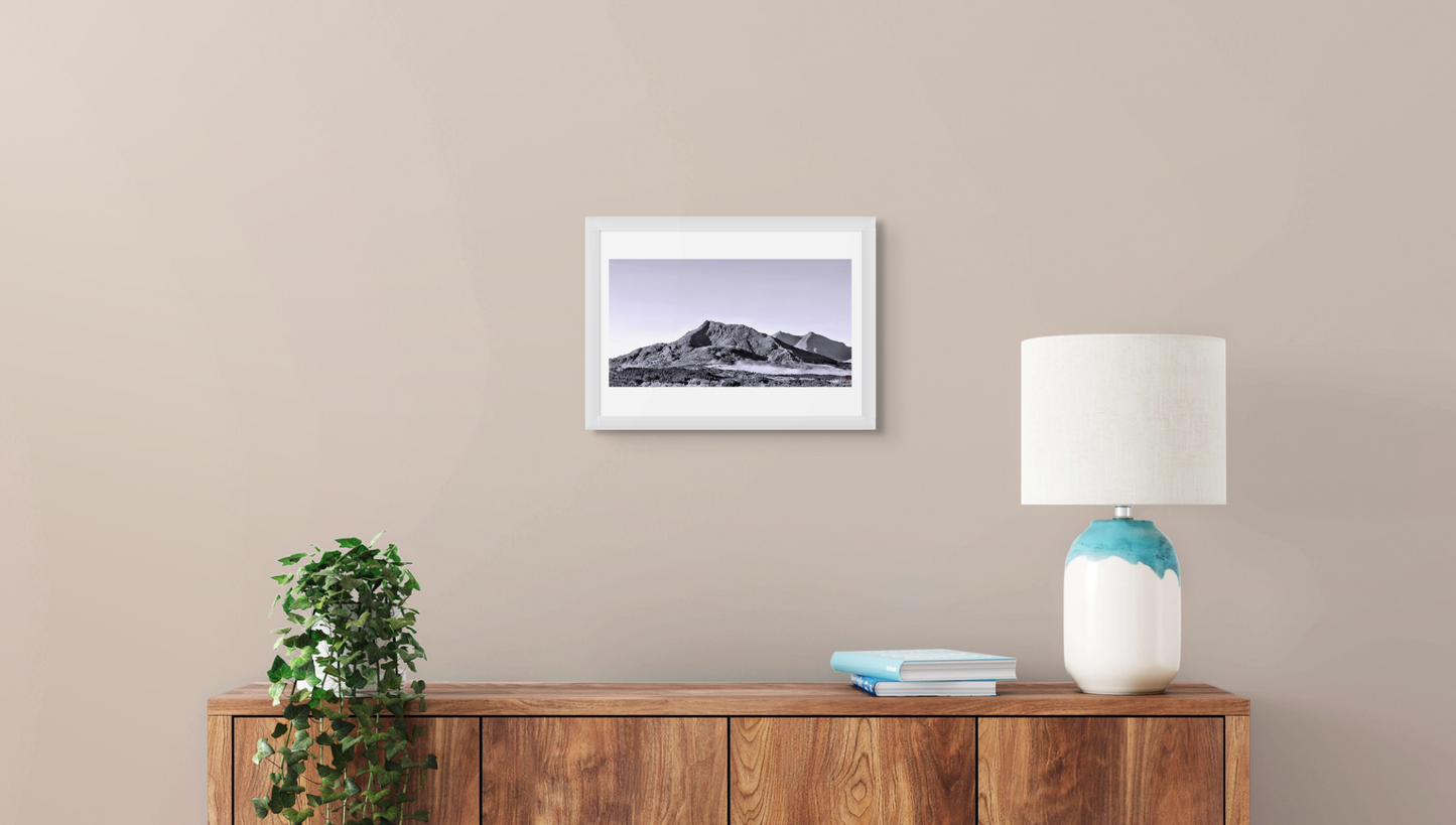 Framed landscape print on a wall above a wooden sideboard with a lamp and plant.