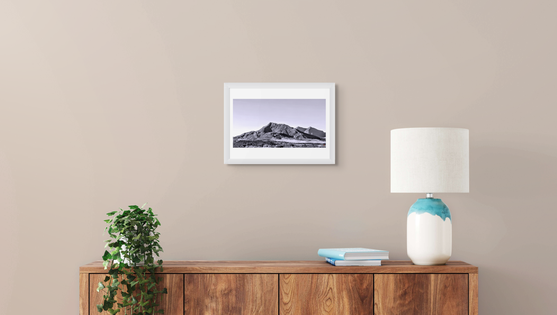 Framed landscape print on a wall above a wooden sideboard with a lamp and plant.