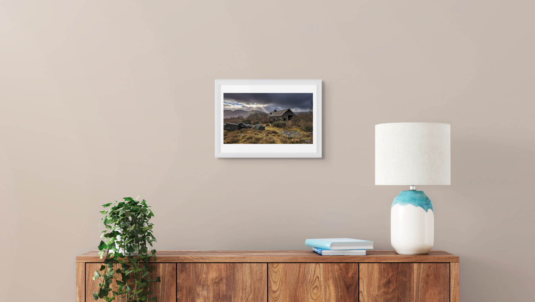 Framed landscape print on a wall above a wooden sideboard with a lamp and books.