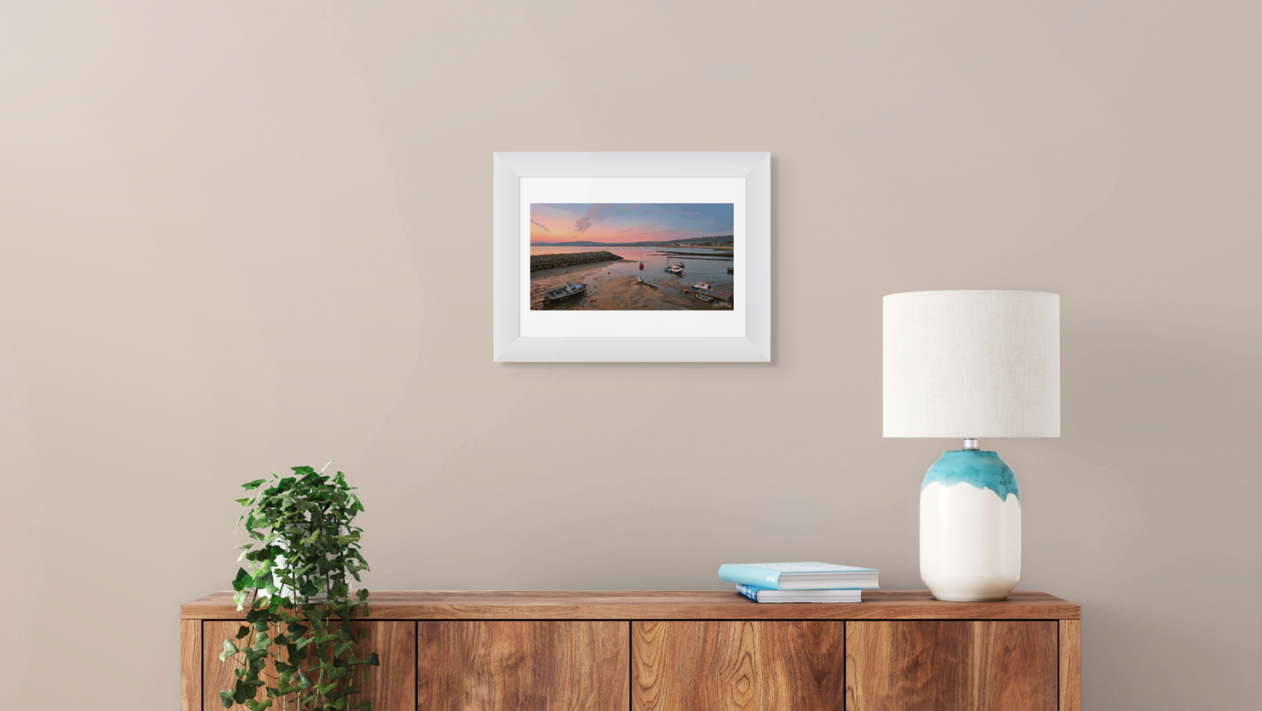 Framed photograph of a sunset over water on a beige wall above a wooden sideboard with a lamp and books.