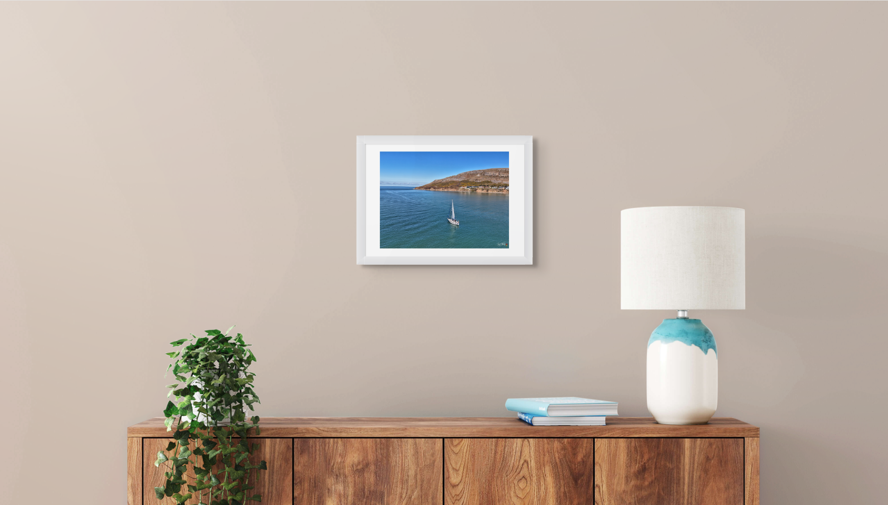 Framed artwork of a sailboat on water above a wooden sideboard with a lamp and plant.