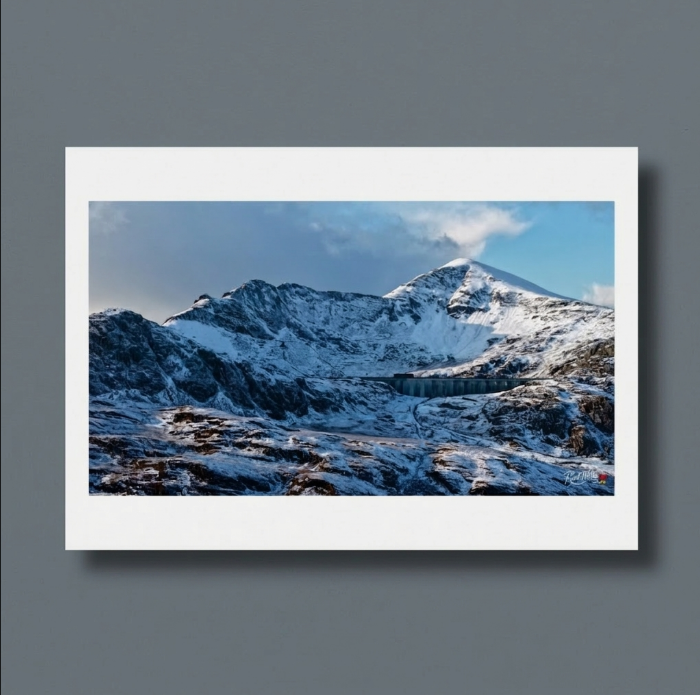 Stwlan Dam is etched into the harsh, snow-covered slopes of the Moelwyns. Long celebrated as a marvel of mid-century hydroelectric ingenuity and a solitary beacon for determined hikers, this remote alpine reservoir retains a deeply formidable, yet industrial and wild, charm. Its sweeping, rhythmic concrete arches and stark, man-made symmetry offer a rigid, resolute contrast to the chaotic, jagged ridges and sheer, snow-filled gullies of the mountains towering immediately above.