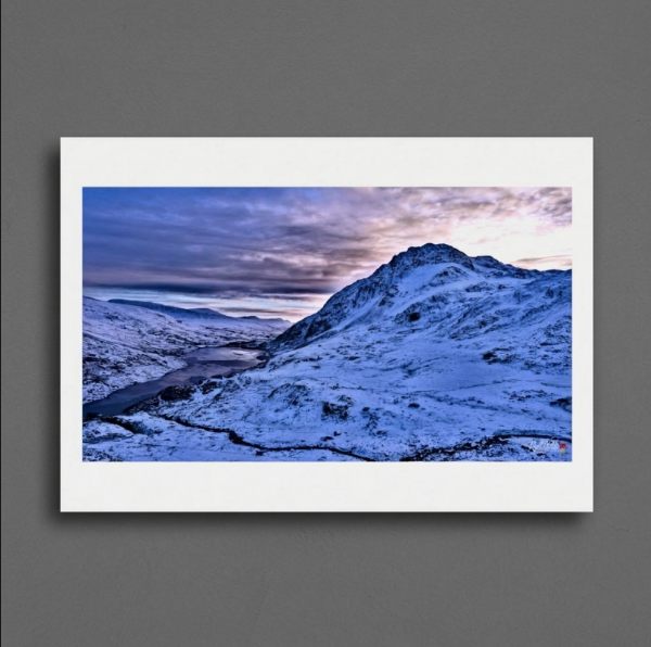 Tryfan rises aggressively into the fading winter light. Long revered as one of Britain's most iconic peaks and a formidable rite of passage for serious scramblers, this jagged monolith retains a deeply wild, unforgiving charm. The dark, still waters of Llyn Ogwen pooling silently at its base offer a flat, sombre contrast to the imposing, heavily fractured ridges and steep, icy flanks towering dramatically above.