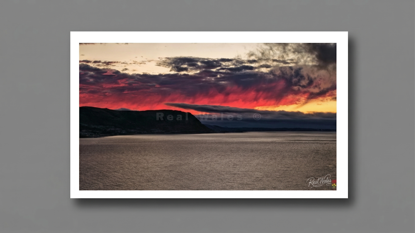 The vibrant sky over West Shore Llandudno, ignites in a breathtaking display of atmospheric fire, as the final light of a winterâ€™s day bleeds across the horizon in a defiant, crimson pulse. Characterised by a singular, horizontal streak of incandescent red and electric magenta, the sky appears to tear open beneath a heavy shroud of bruised, charcoal clouds. This low-slung belt of light casts a haunting, vertical shimmer resembling a phantom rain of colour that spills toward the dark, silhouette-etched pro
