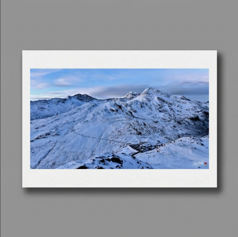 The vast, snow-draped massif of Yr Wyddfa rises dramatically into the winter sky. Long revered by adventurous mountaineers, this formidable landscape retains a deeply wild, majestic charm. The delicate, winding mountain paths traced into the pristine white snow and the small, clustered roofs of the pass below offer a quiet, humble contrast to the imposing, jagged ridges and sheer, frozen faces of the highest peak in Wales.